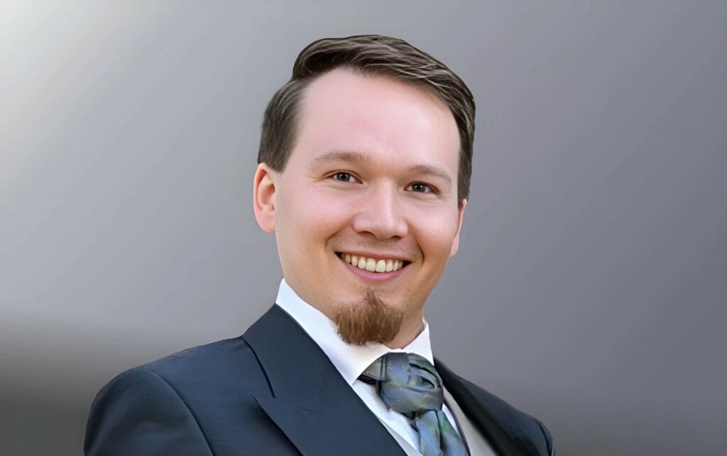 Foto_RobertSkawinski Porträt von Robert Skawinski, ein lächelnder Mann im Anzug mit Krawatte vor neutralem Hintergrund.