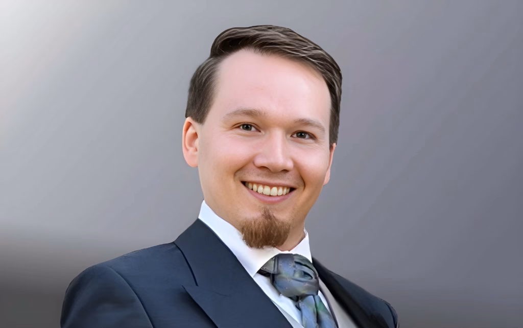 Foto_RobertSkawinski Porträt von Robert Skawinski, ein lächelnder Mann im Anzug mit Krawatte vor neutralem Hintergrund.