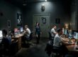 Dunkler Raum mit mehreren Personen an Computern, eine Frau steht in der Mitte, an den Wänden hängen düstere Porträts und ein Totenkopf.