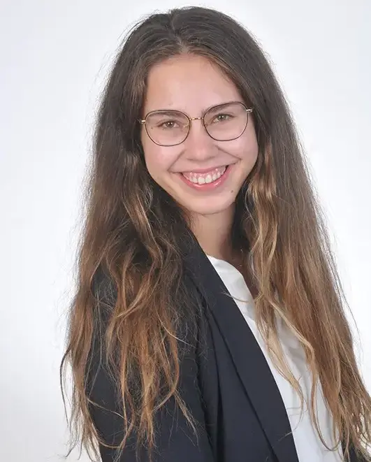 Daniela_Soeldner Porträt von Daniela Söldner, eine lächelnde junge Frau mit langen welligen Haaren und feiner Brille, im dunkelblauen Blazer über weißem Oberteil vor hellem, neutralem Hintergrund.